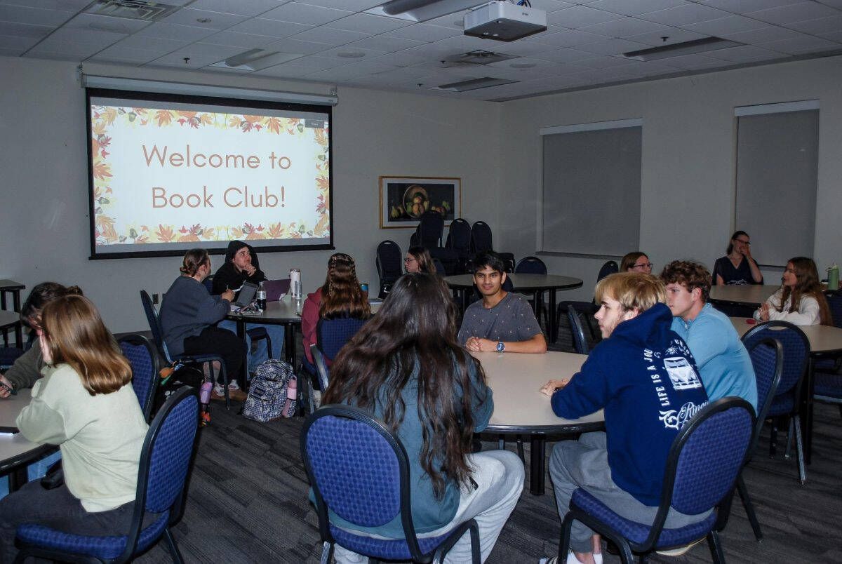 Grand Valley Students sitting in a low lit room with a projector that reads 'Welcome to Book Club!'